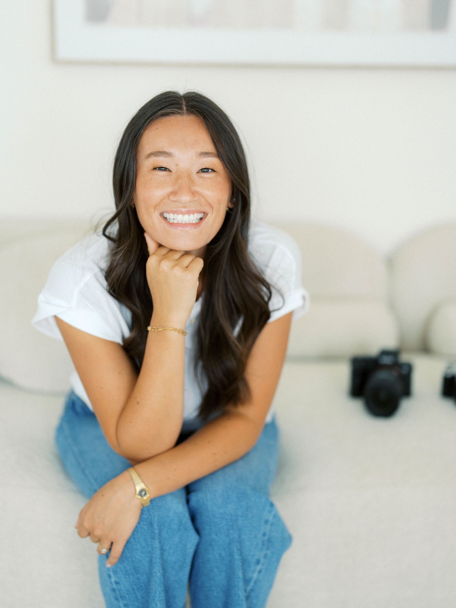 Smiling young woman in white t-shirt and jeans sitting on a couch with a camera in the background.