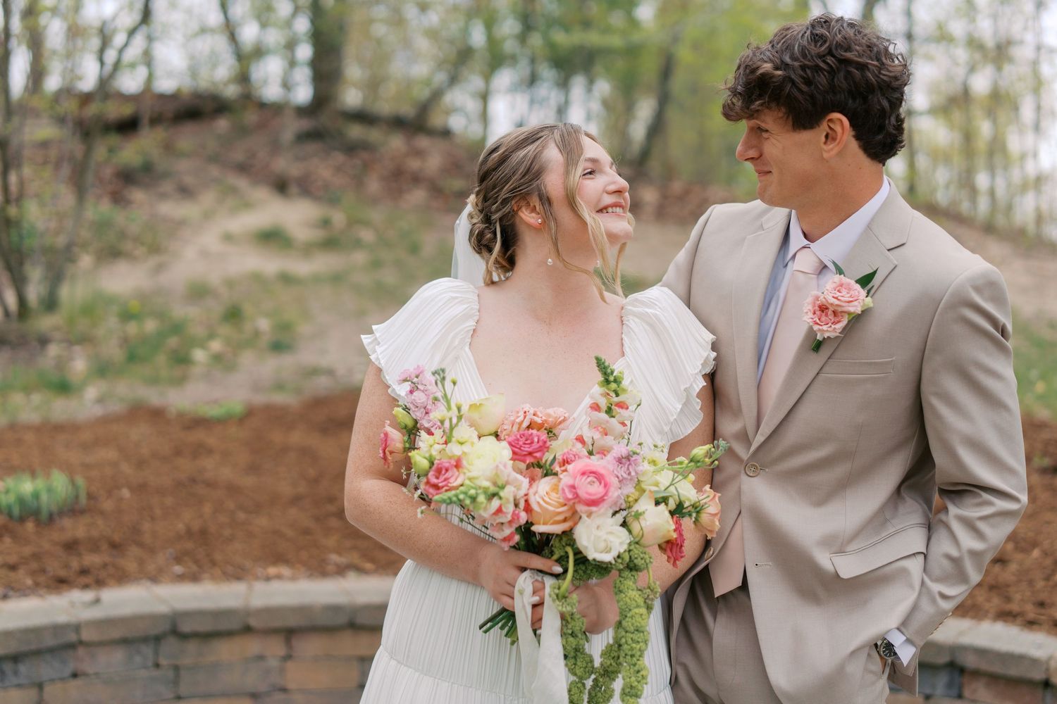 Bride and groom smiling at each other outdoors, bride holding colorful spring bouquet, groom in tan suit with floral boutonniere.
