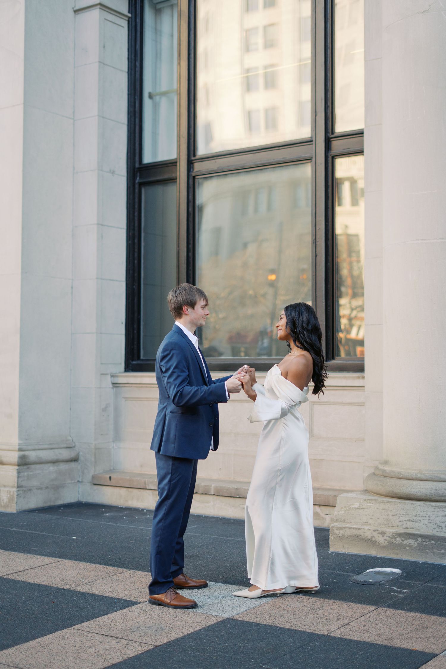 A couple in white dress and navy suit dances playfully outside a building.