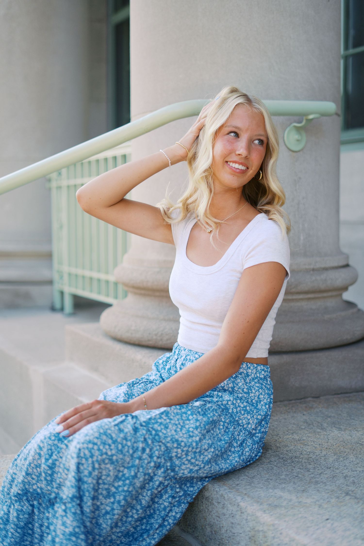 A person in a white top and blue floral skirt poses gracefully against a stone column backdrop.