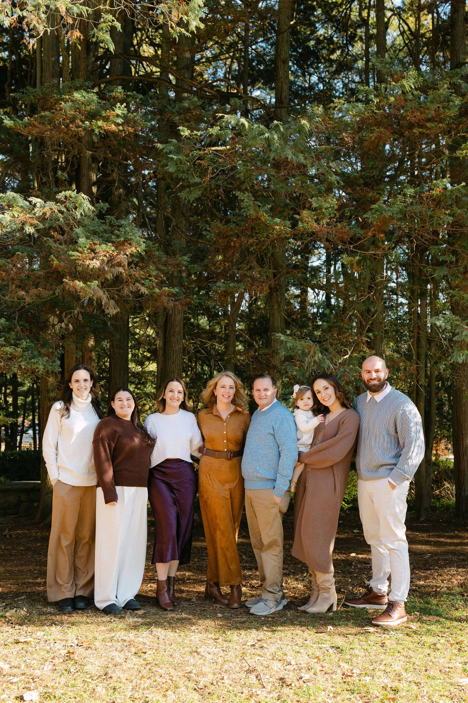 Group of people in earth-toned clothing stand together outdoors in front of autumn trees.