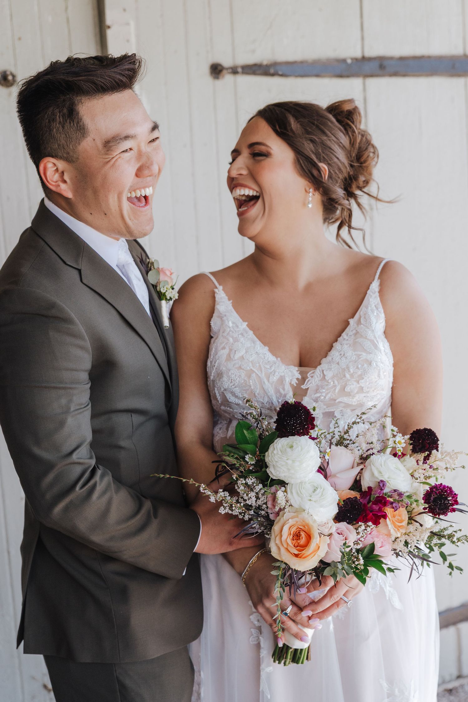 A happy couple shares candid laughter during their wedding photoshoot while holding a colorful floral bouquet.