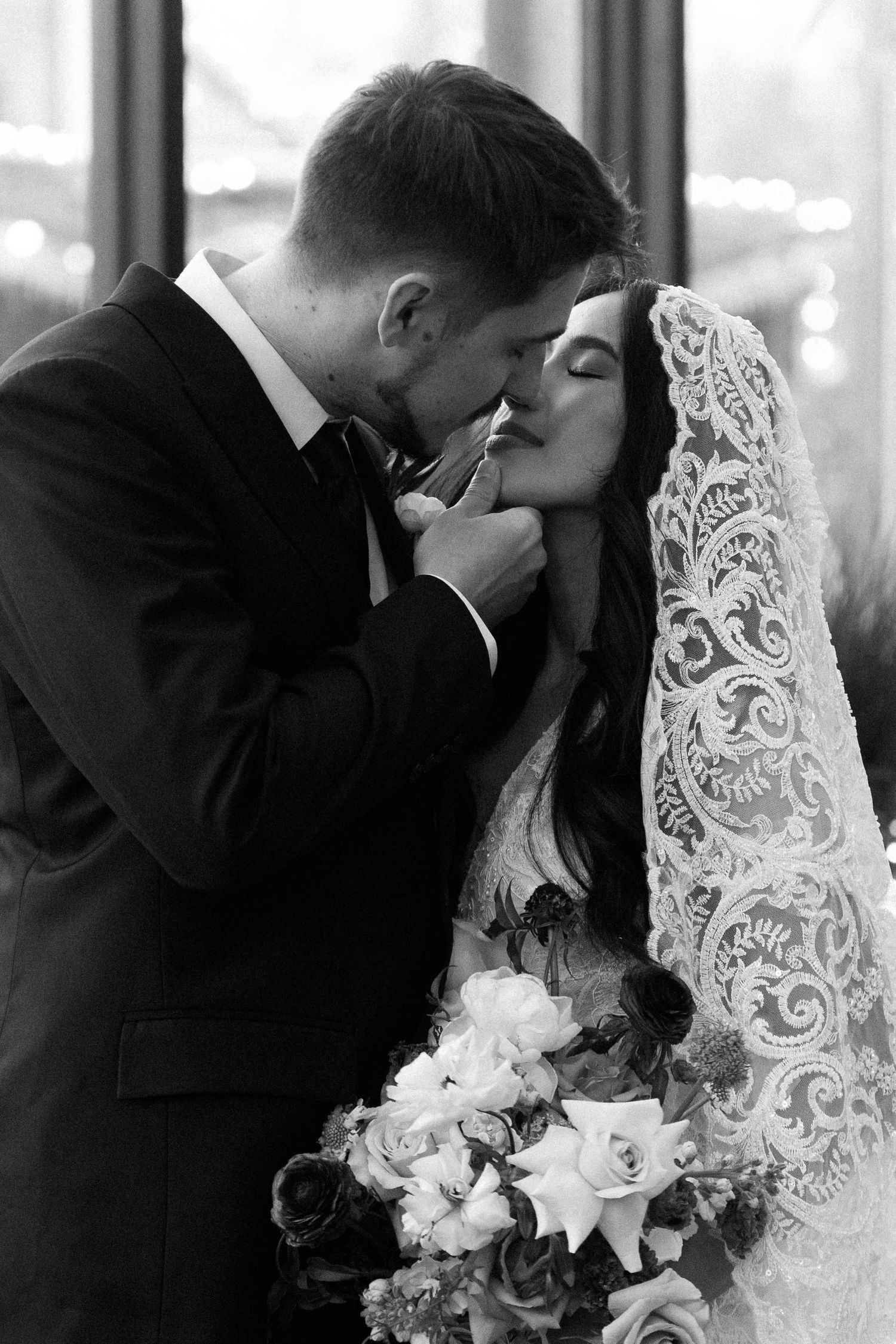 A romantic black and white wedding photo showing a tender moment between newlyweds with a lace veil and floral bouquet.