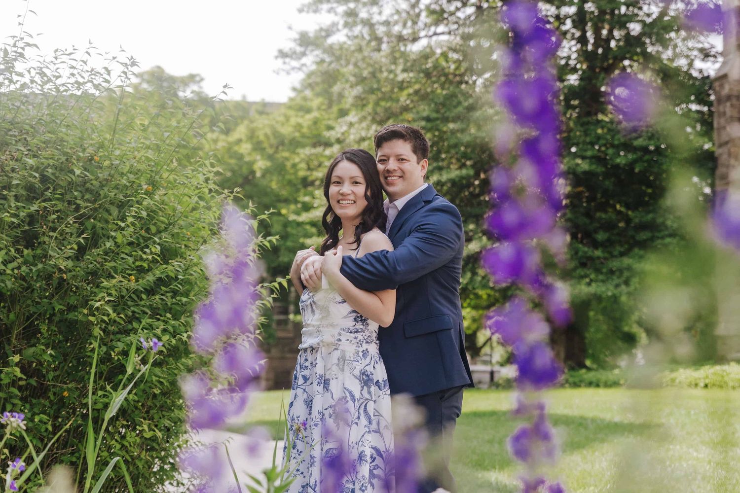 A couple shares a romantic moment among blooming purple delphinium flowers in a lush garden setting.