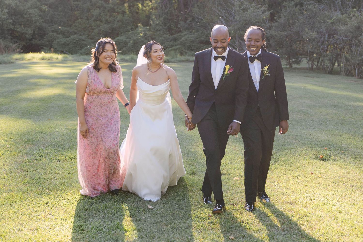 Wedding party members walk together on a sunlit grassy lawn during an outdoor celebration.