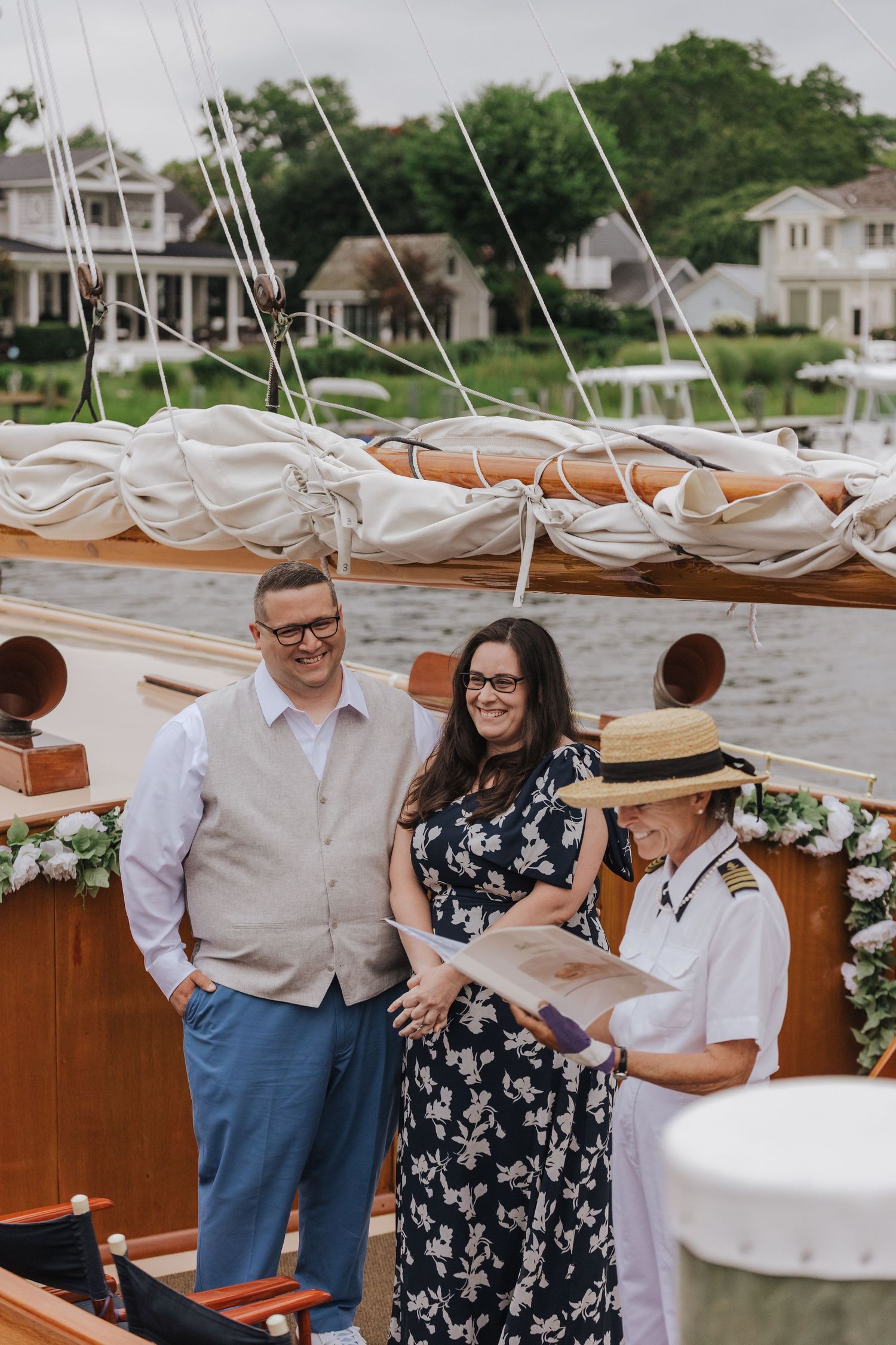 A couple stands together on a boat deck with water and houses visible in the background on a sunny day.