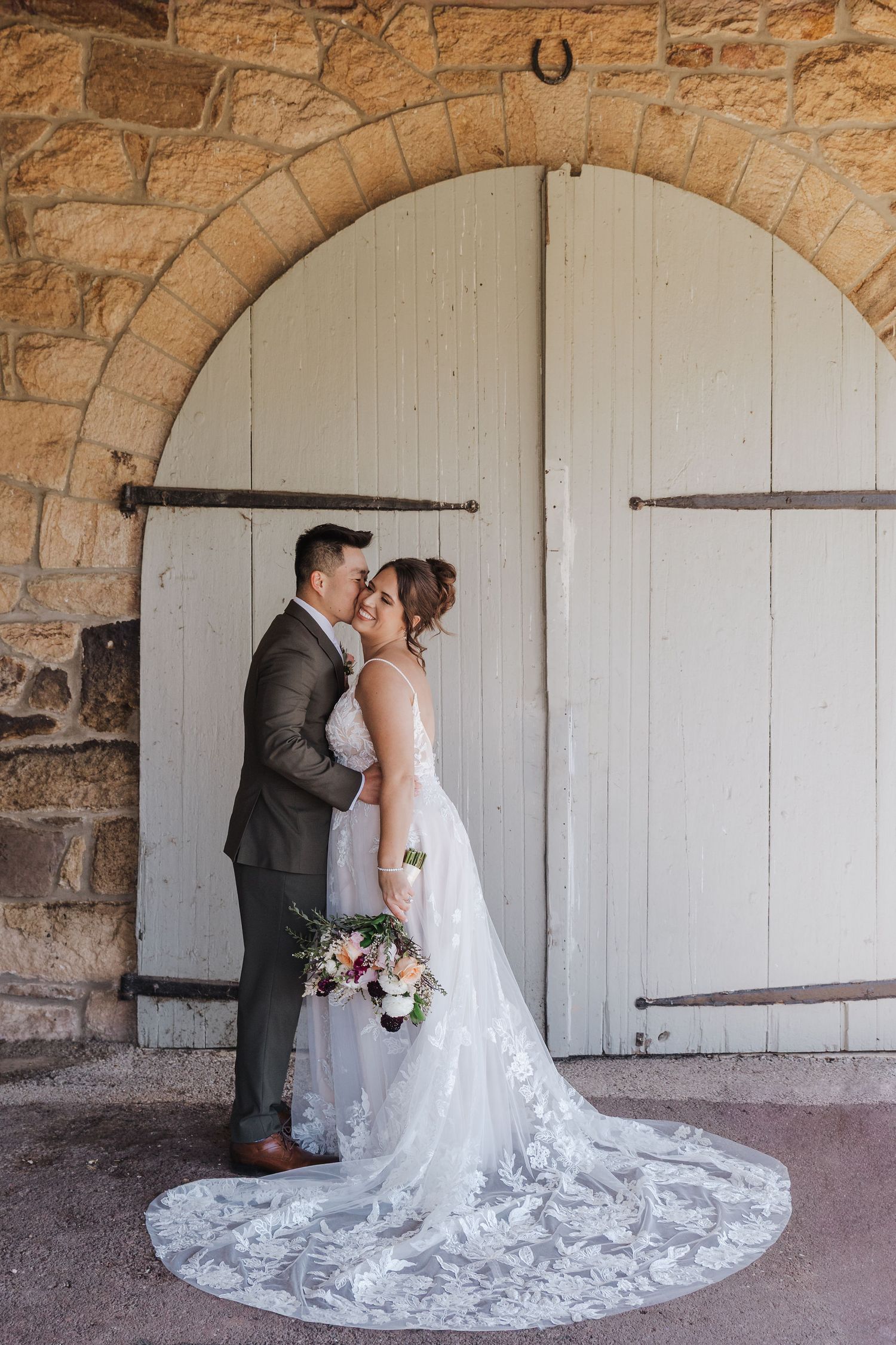 A romantic wedding couple shares intimate moments against a rustic stone wall and white barn door backdrop.