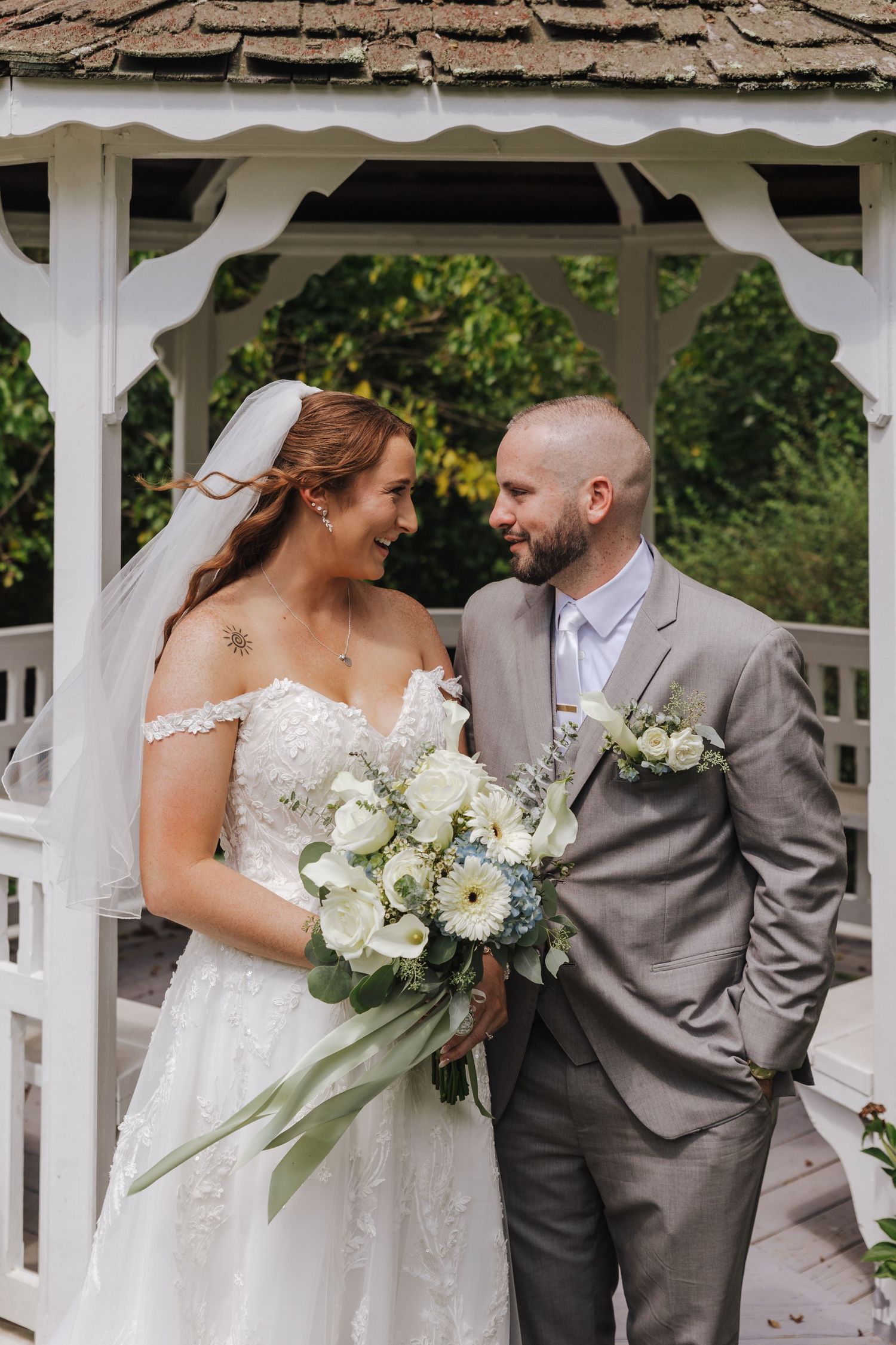 A couple shares a joyful moment together under a white gazebo while holding elegant white floral bouquets.