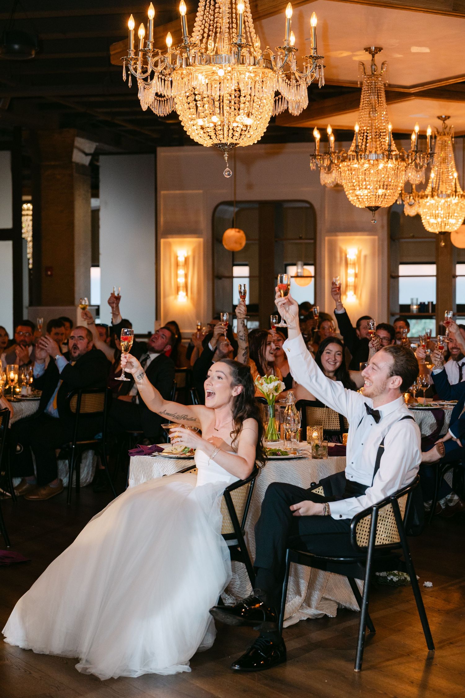 Wedding couple and guests perform a fun synchronized dance move during reception under crystal chandeliers.