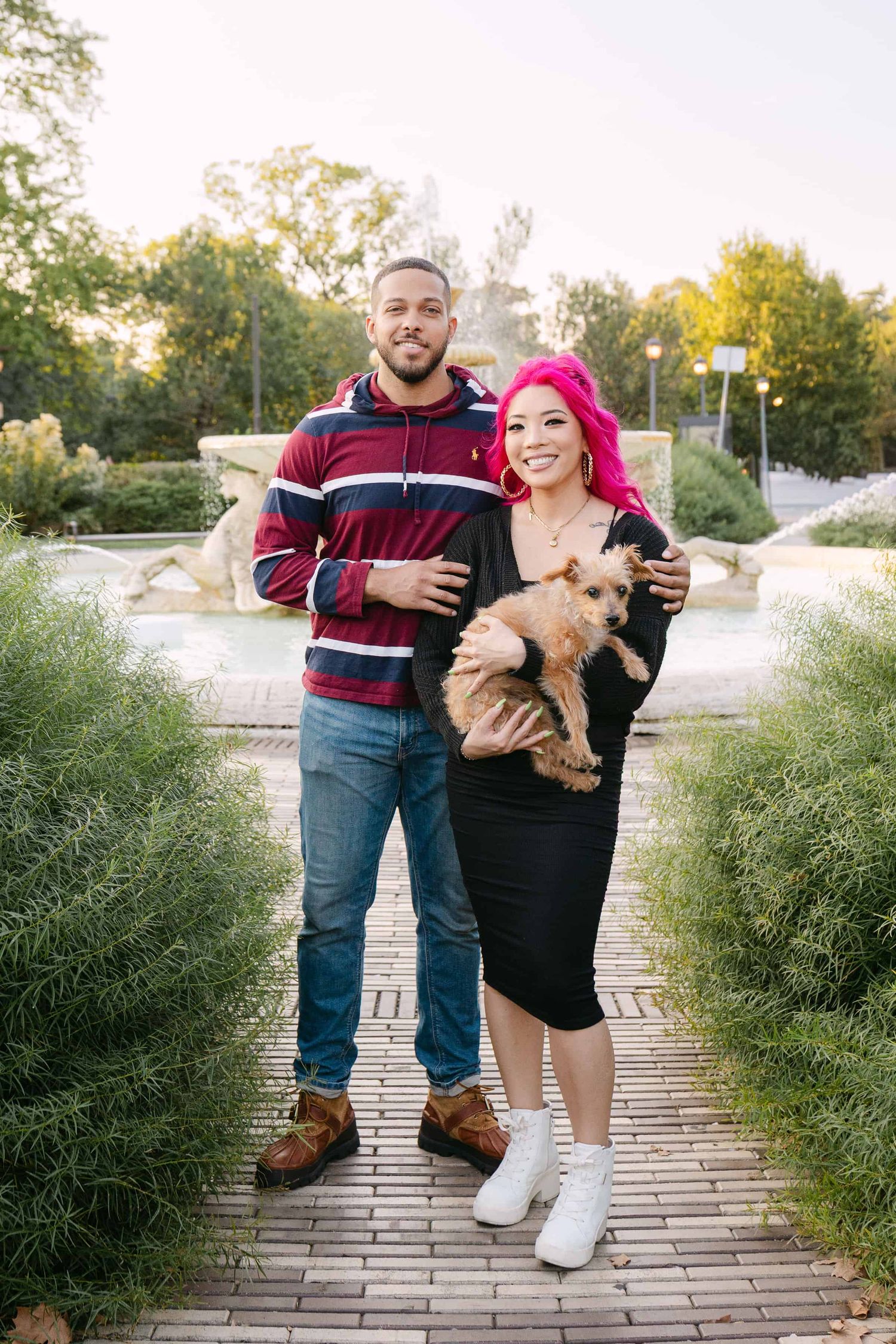 A couple poses with their small dog on a brick pathway surrounded by green bushes during golden hour.