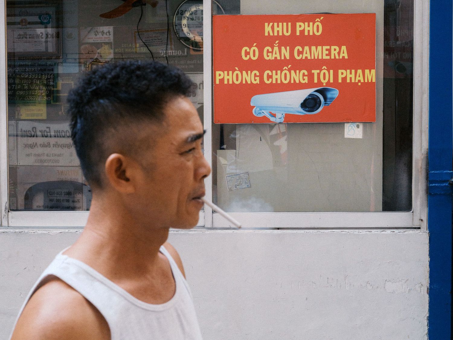 Profile of a man in a white tank top walking past a red Vietnamese sign warning that security cameras are installed to prevent crime.