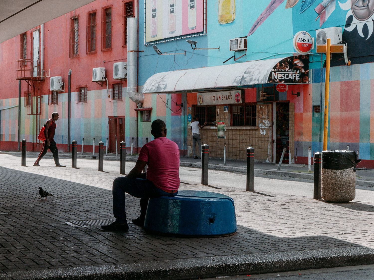 Man in a red shirt sits on a blue stool on a sunlit city sidewalk beside colorful street art buildings, with a pedestrian walking past.