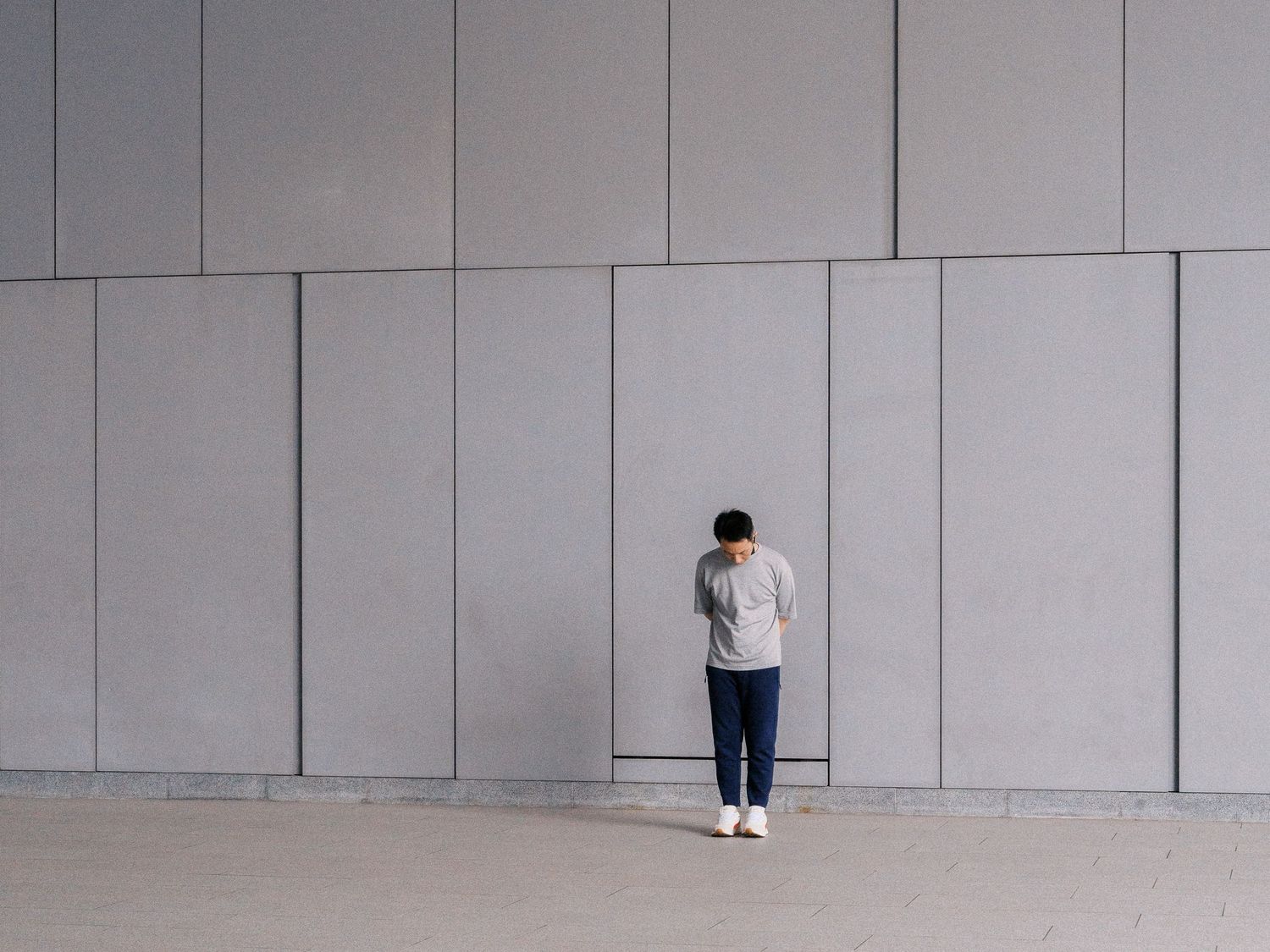 Person in a gray sweater and dark pants bows their head while standing alone before a large minimalist gray paneled wall.