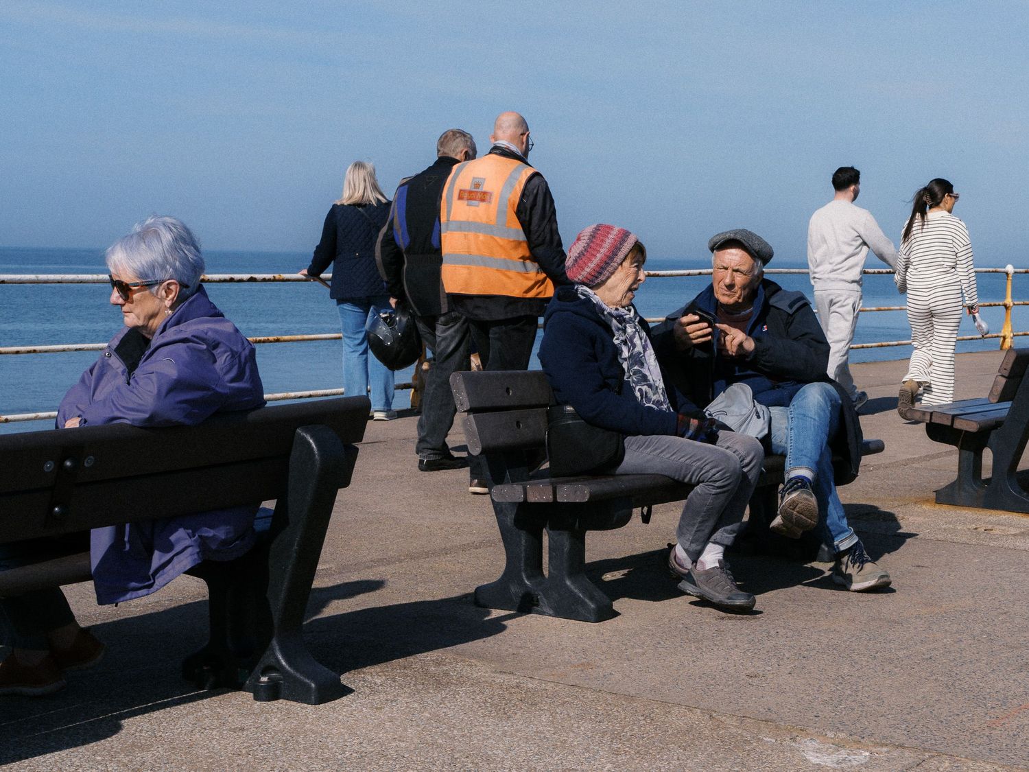People relax on wooden benches along a sunny seaside promenade while others stroll past the railing overlooking calm blue water.
