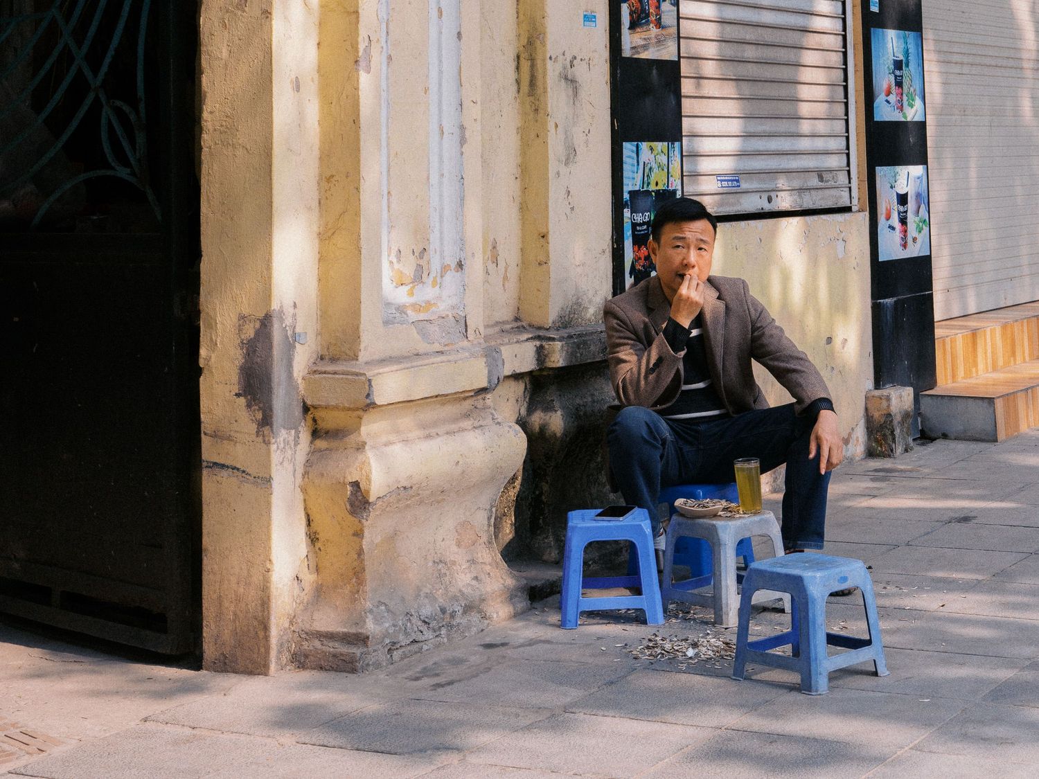 Man in a gray blazer sits on a small blue plastic stool beside a weathered building, smoking on a quiet street with a drink nearby.