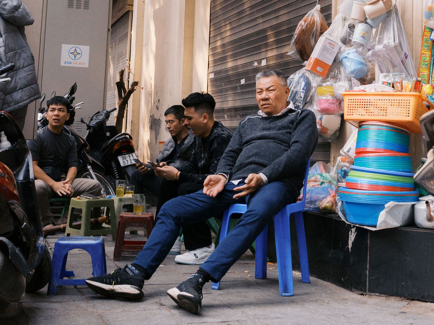 Men sit on small plastic stools along a busy street sidewalk beside stacked colorful plastic basins and parked motorbikes.