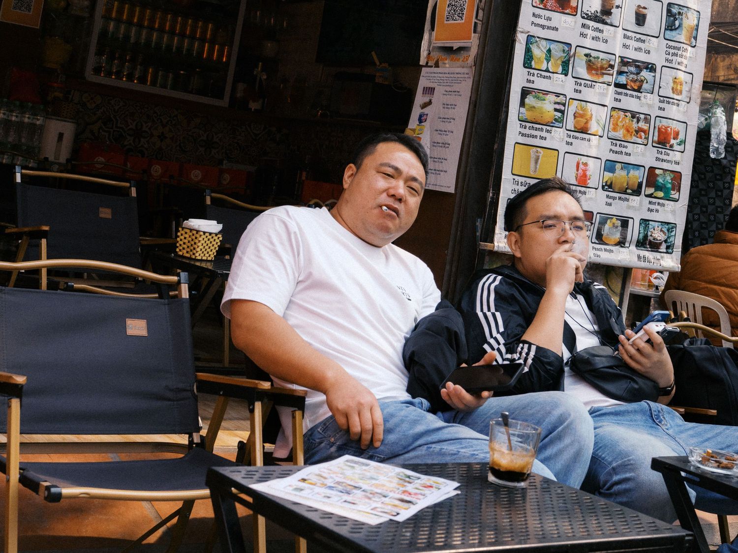 Two men relax at an outdoor café table with iced coffee, one in a white t-shirt and one smoking, beside a menu poster board.
