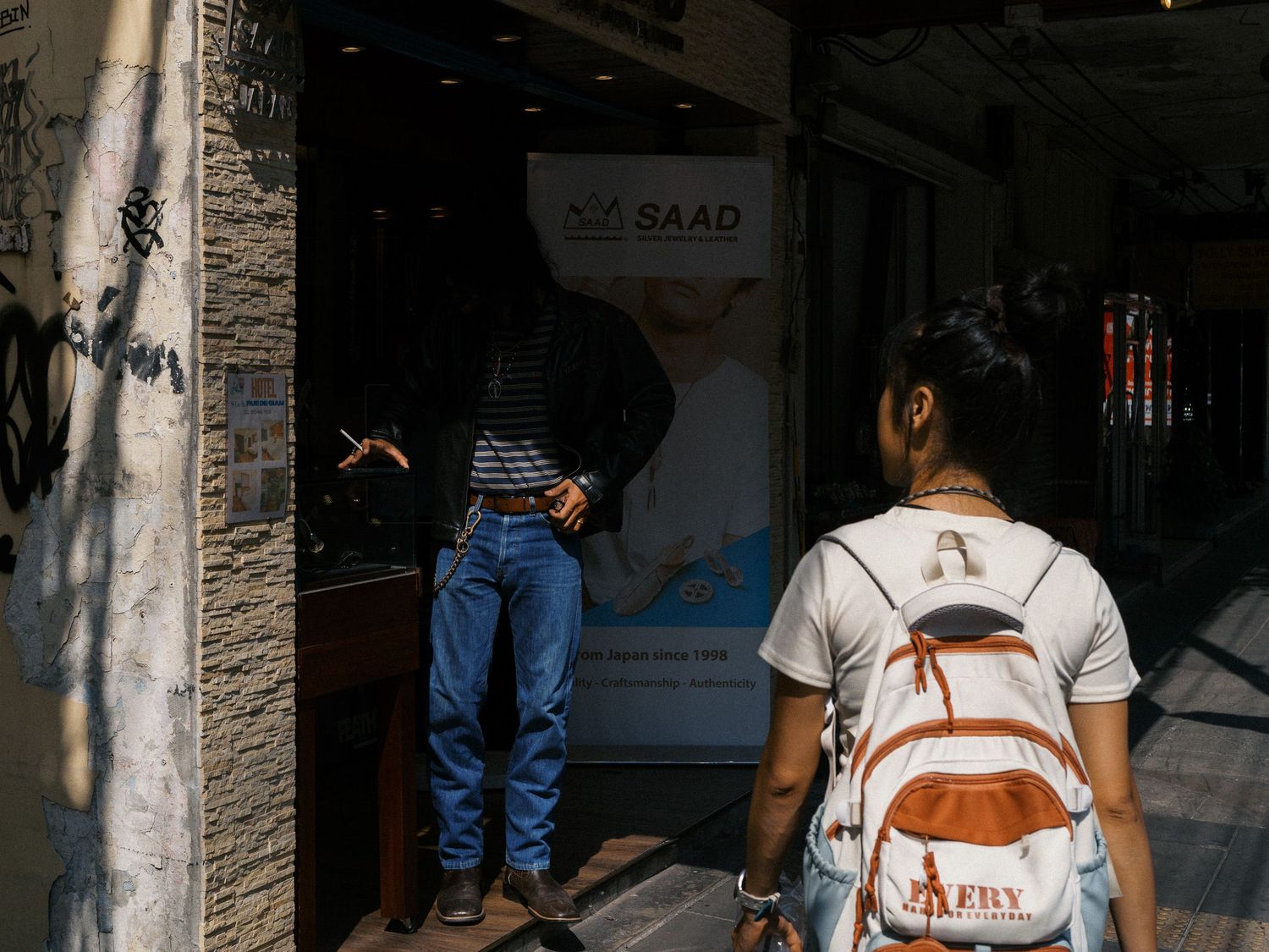 Woman with an orange and white backpack walks past a man in jeans standing outside a shop with a SAAD poster, near a graffiti-covered wall.