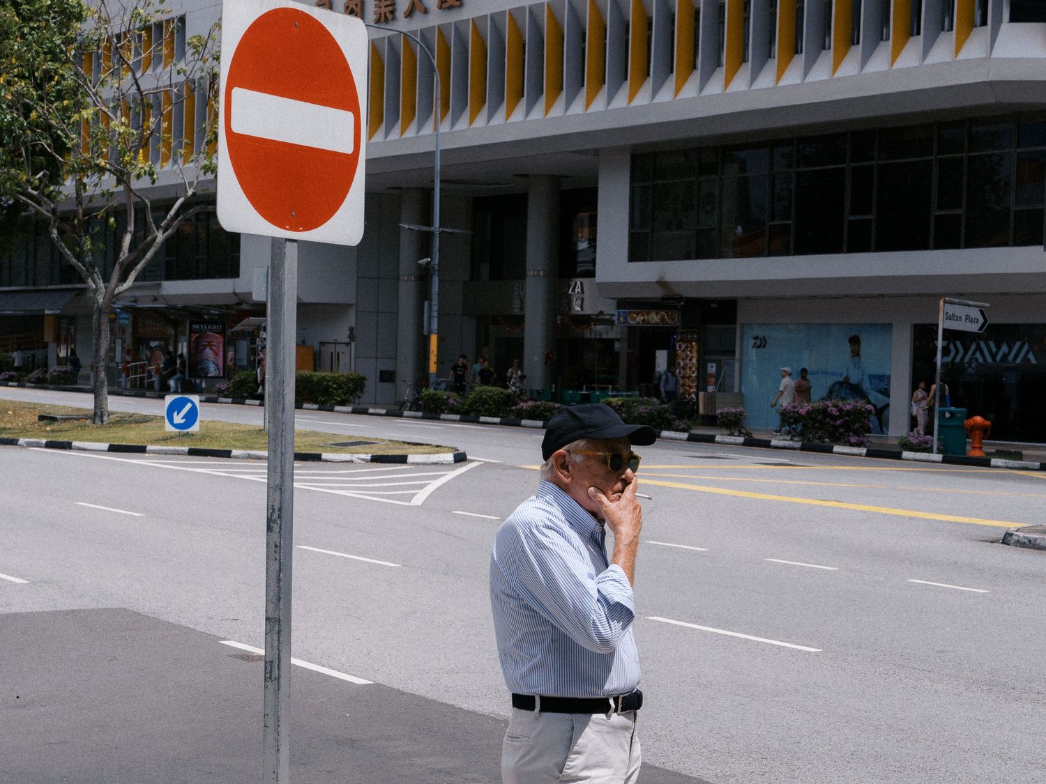 Man in a light blue shirt and cap stands beside a red no-entry sign on an empty city street, with a modern building behind him.