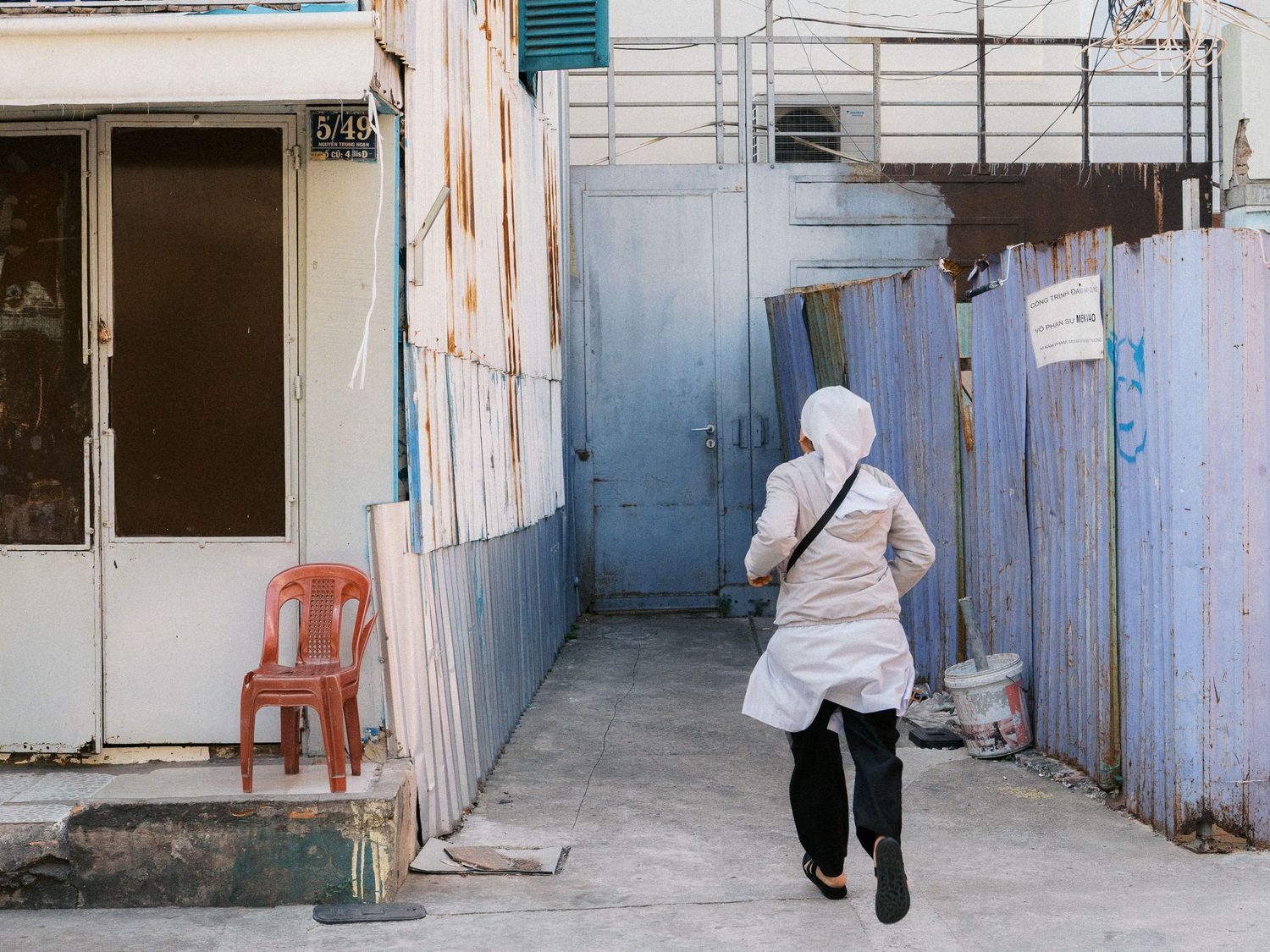 Woman in a white headscarf and apron walks down a narrow alley past a red plastic chair and blue corrugated metal fences.