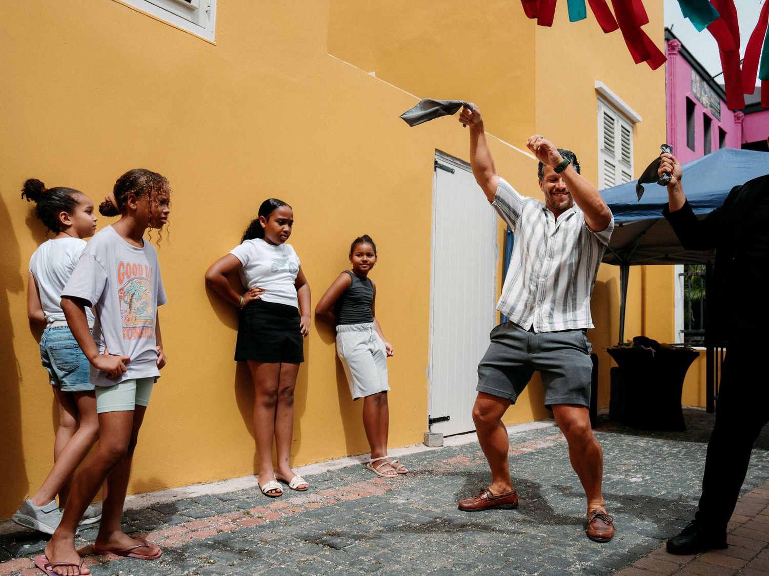 Young girls watch a smiling man in striped shirt and shorts dancing joyfully on a cobblestone street beside a bright yellow building.