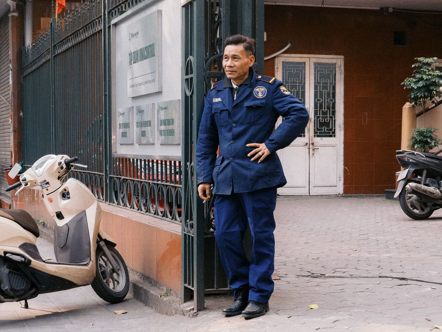 Security guard in a navy blue uniform standing with hand on hip beside a black metal gate, with parked scooters on a paved street.