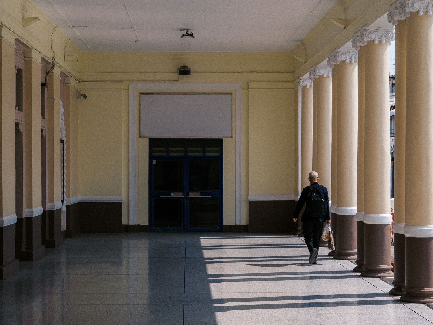 Person walking along a sunlit colonnaded corridor with cream-colored columns, polished marble floor, and dark double doors at the end.