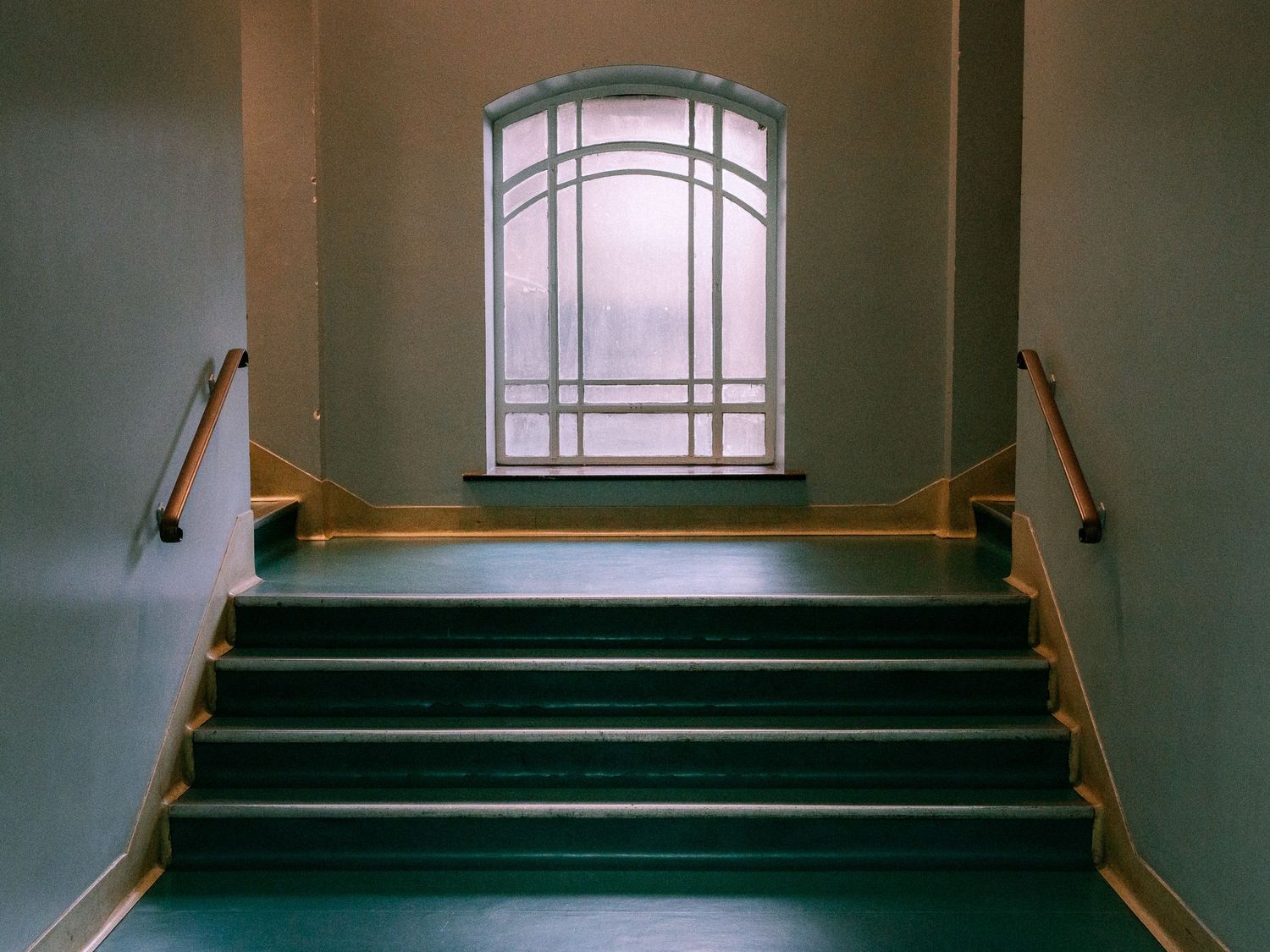 Wide green staircase with wooden handrails leading up to a large arched window with frosted glass in a softly lit, empty interior hallway.