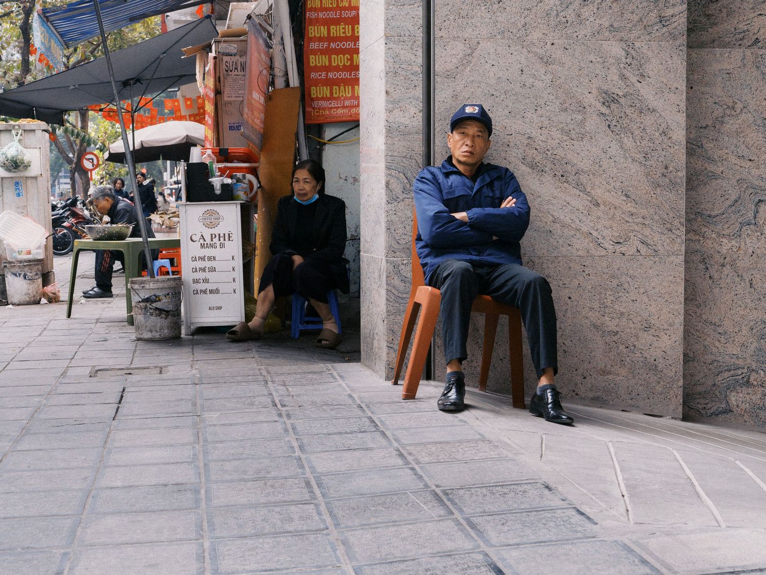 Man in a blue uniform and cap sits with arms crossed on a wooden stool along a tiled sidewalk beside a street vendor's stall.