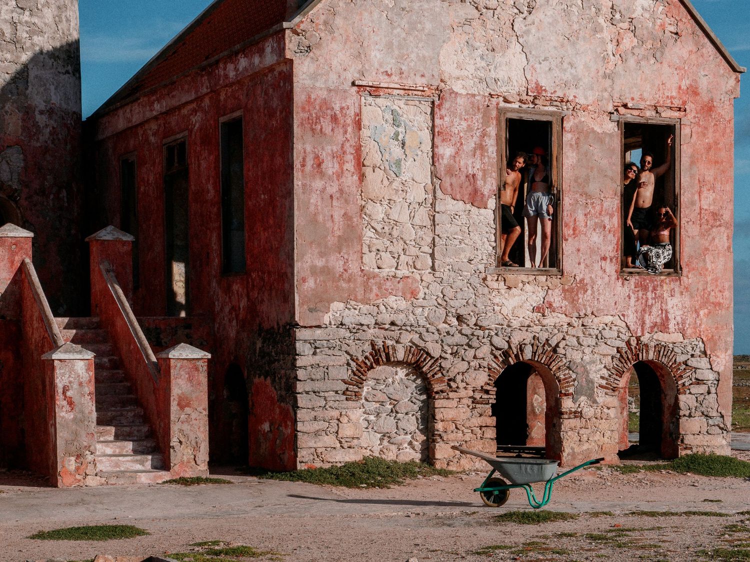 Weathered pink colonial building with stone arches and exterior staircase, people standing in upper windows and a green wheelbarrow in the f