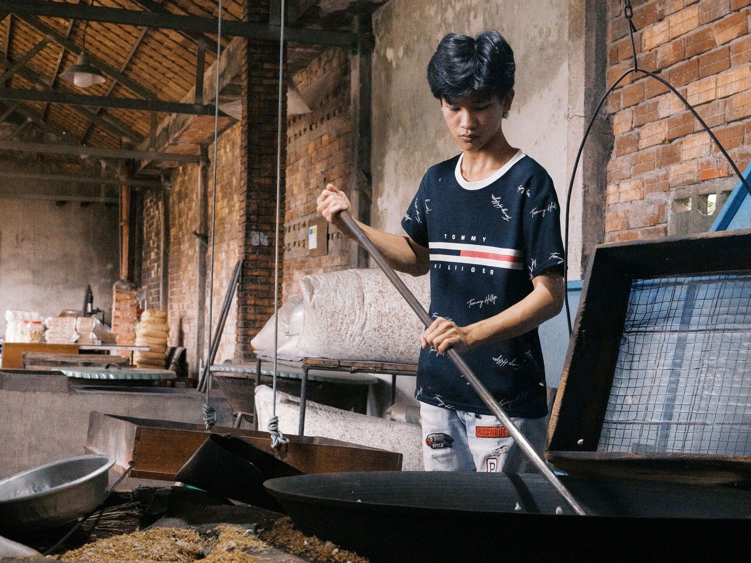 Young man in a dark striped shirt stirs a large wok with a long wooden paddle in a rustic brick workshop.