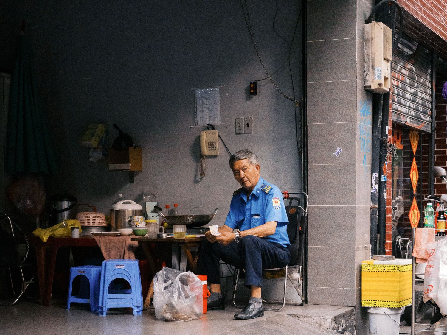 Uniformed man in a blue shirt sits on a chair beside a small table of kitchenware in a shaded urban alley.