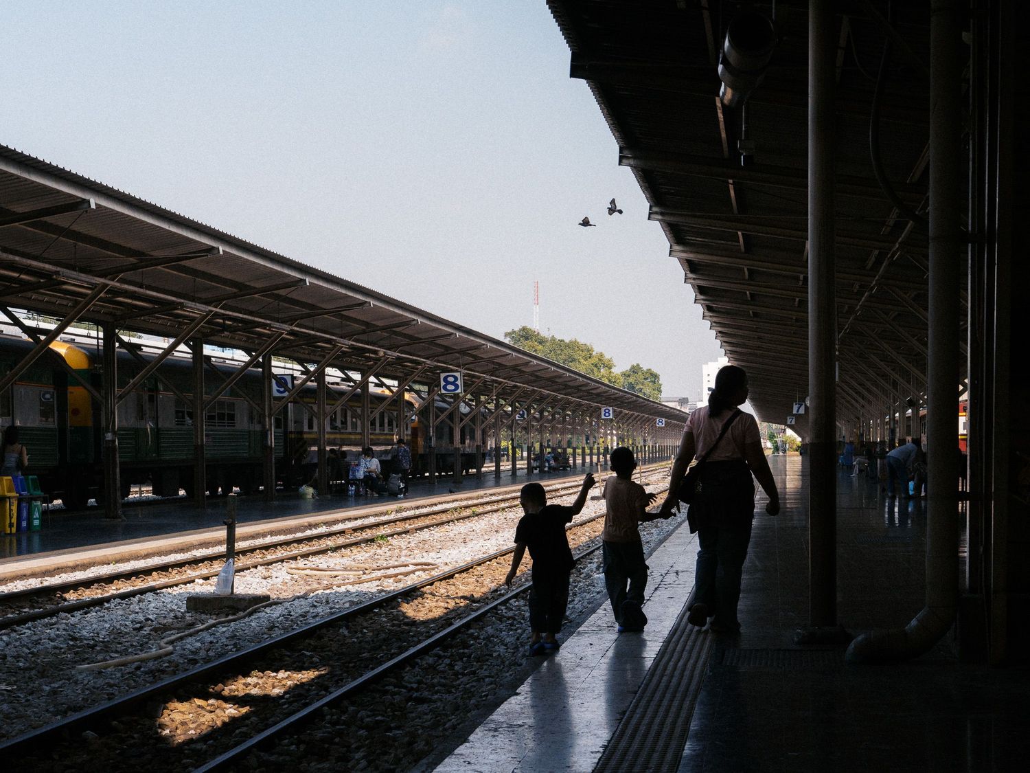 Silhouetted woman and two children walk along a covered train station platform beside parked trains under a bright afternoon sky.