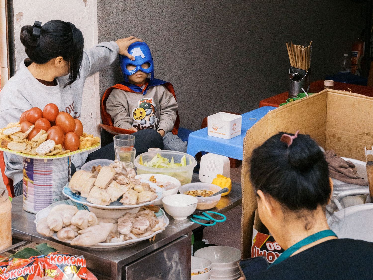 Child in a blue luchador-style mask sits near a street food stall as a vendor arranges plates of steamed buns, sausages and snacks.