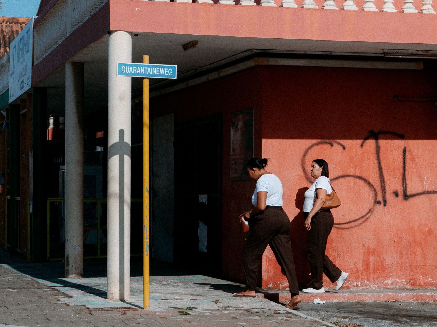Two women in white tops and dark pants walk past a red graffiti-covered building on a sunlit street corner with a blue street sign.