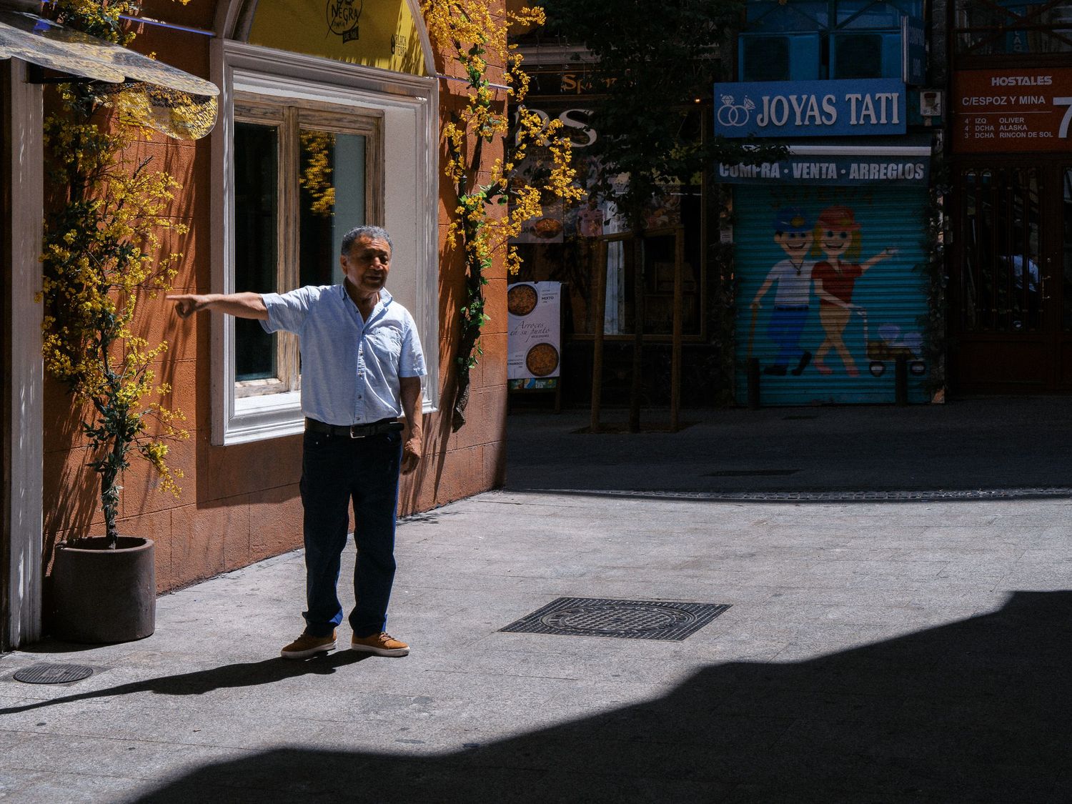 Man in a light blue shirt and dark pants stands on a sunlit pedestrian street beside a flower-covered wall, with shop signs behind him.