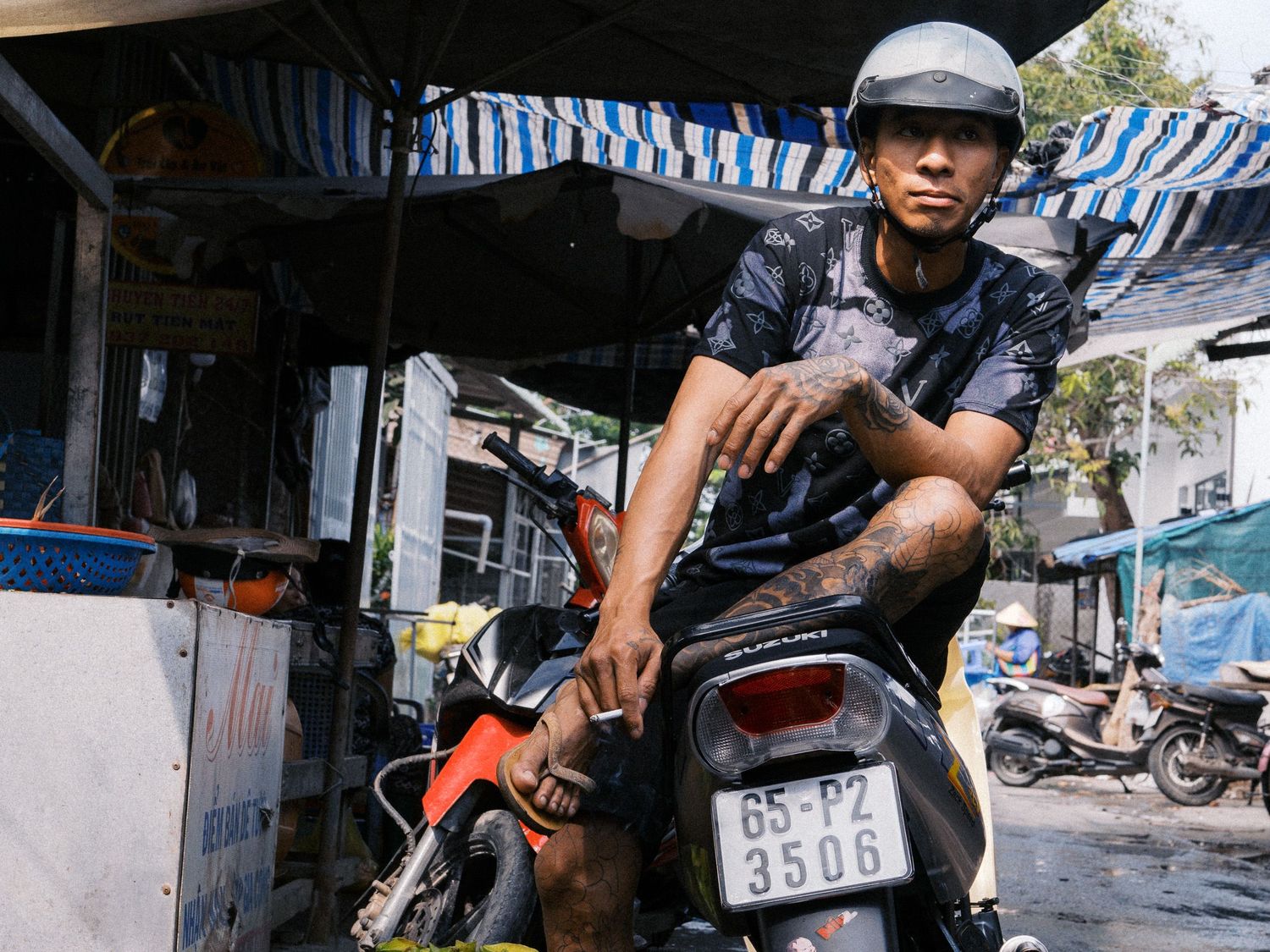 Man in a gray helmet and tattooed arms sits on a motorbike with license plate 65-S2 3506 under a striped awning at a roadside market.