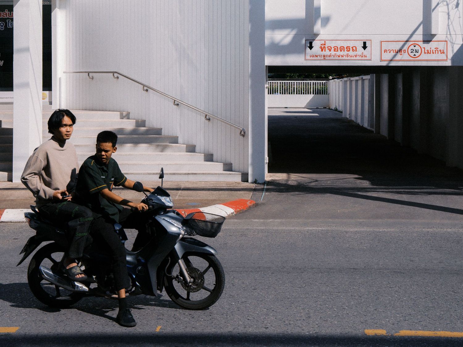 Two young men ride a black motorcycle along a sunlit city street past a white concrete staircase and an underpass with Thai signage.