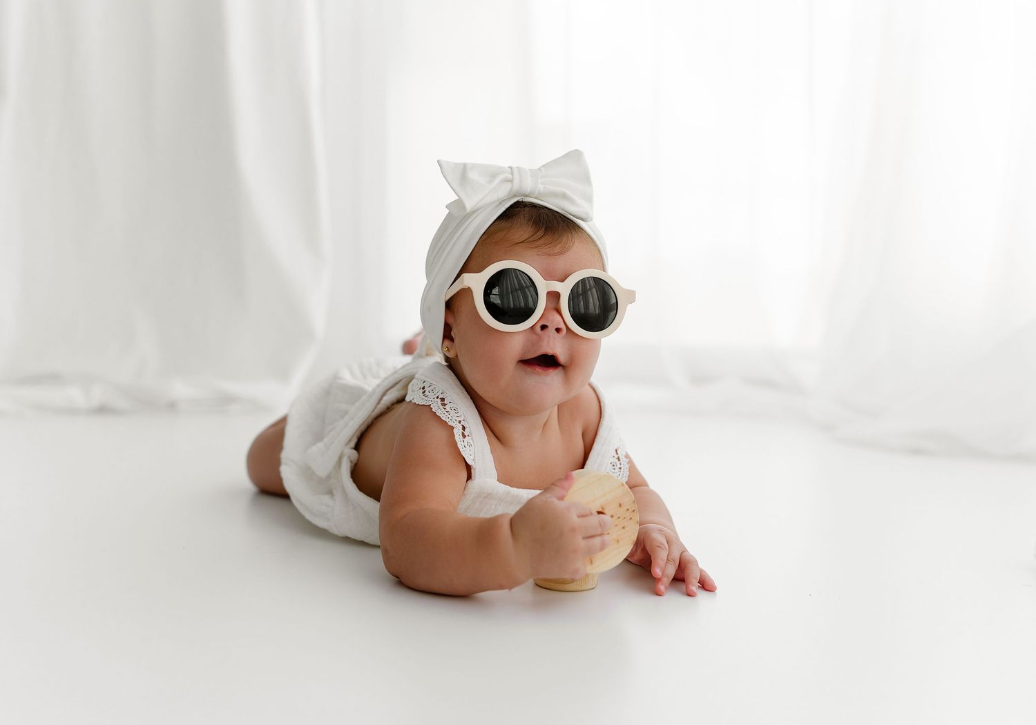 Baby wearing sunglasses and white outfit lying on white background.