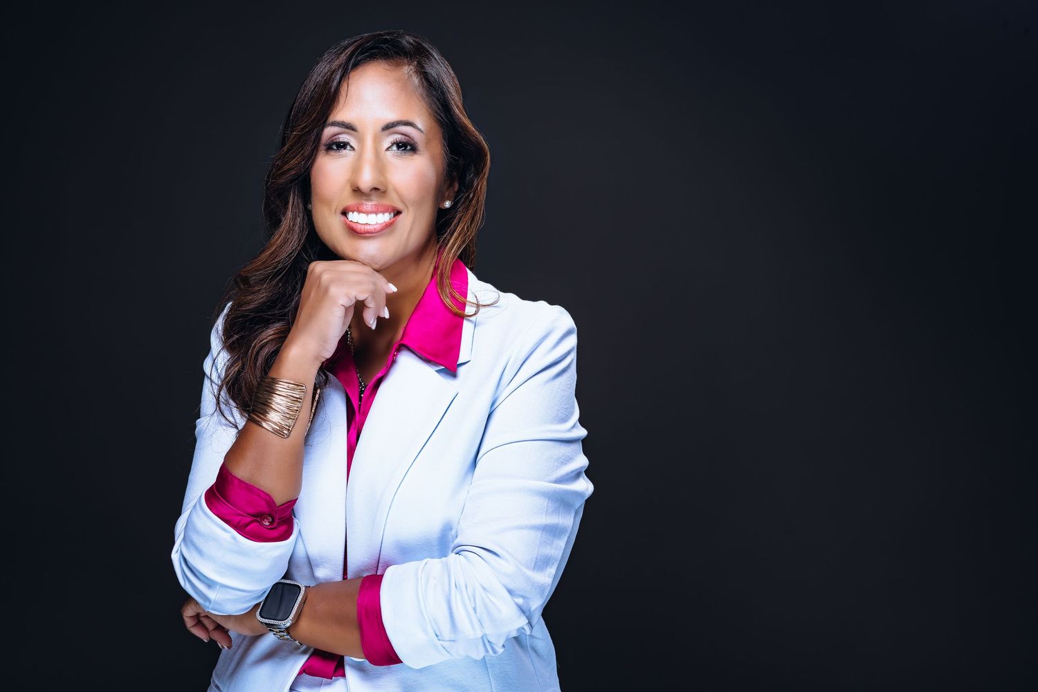 Professional woman in white blazer and pink shirt smiling confidently against dark background.