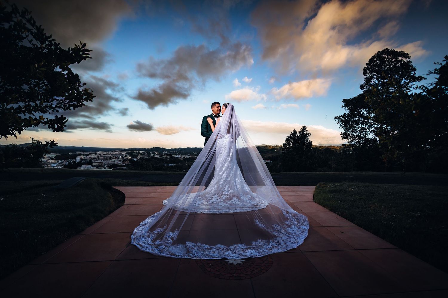 A couple in wedding attire stands on a terrace at sunset with a dramatic lace veil flowing behind them.