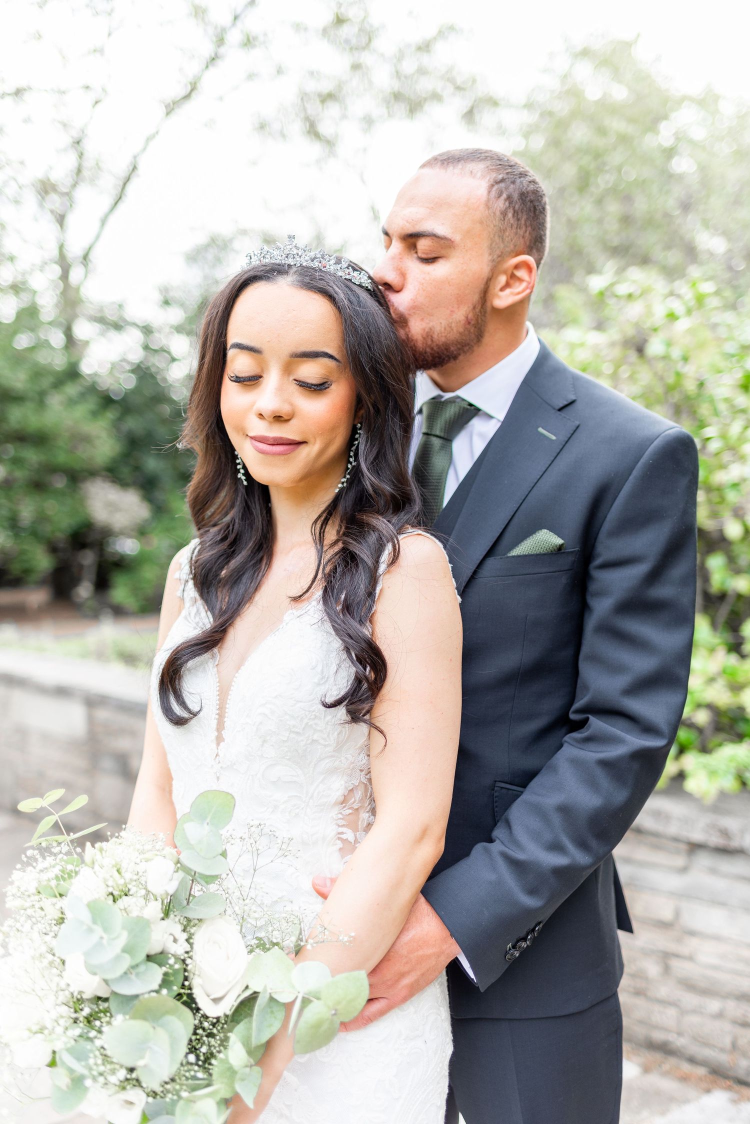 Bride in white lace gown with tiara holding eucalyptus bouquet embraces groom in dark suit during outdoor wedding portrait.