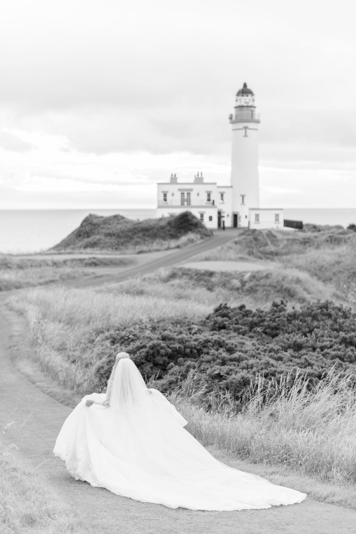 A white wedding dress with a long train trails along a winding path leading to a coastal lighthouse on a cloudy day. Trump Turnberry.
