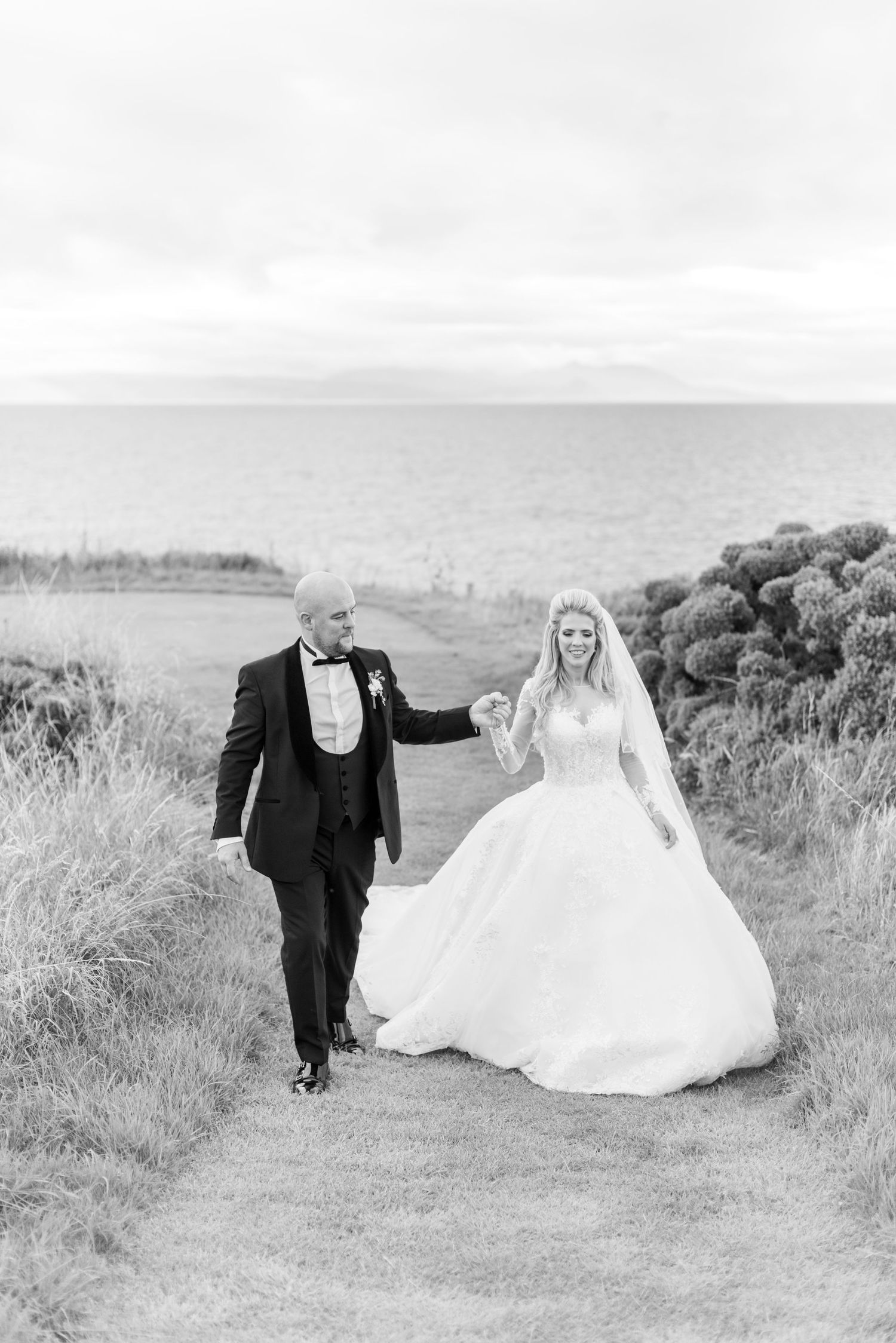 A couple in wedding attire walks together along a scenic coastal path during their black and white outdoor photoshoot. Trump Turnberry.