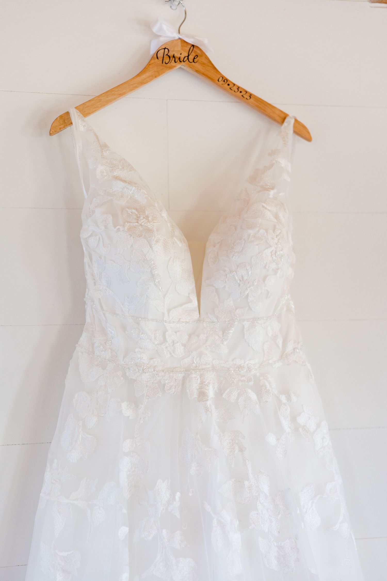 A lace wedding dress with deep v-neckline hanging on wooden bridal hanger against white wall.