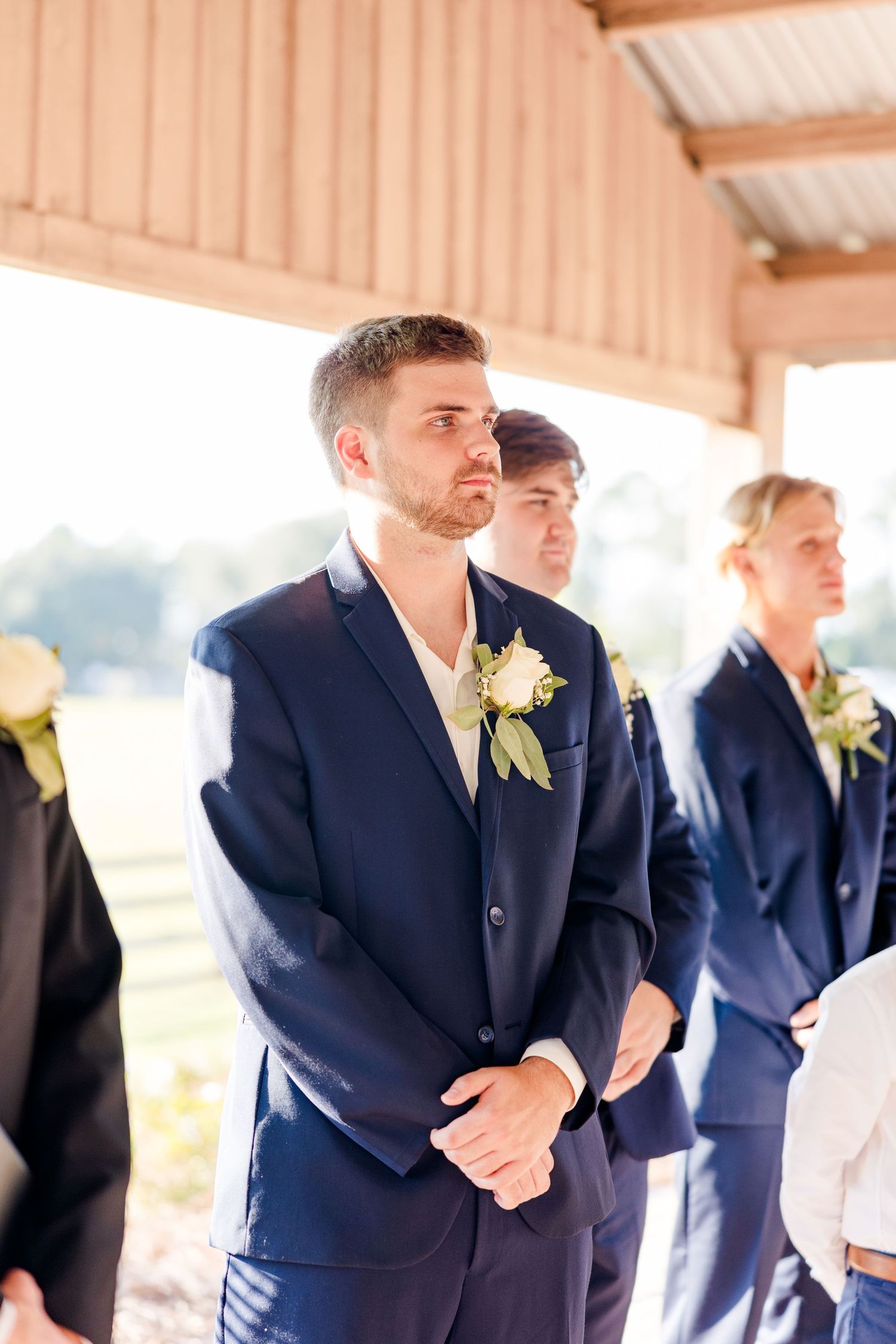 Groomsmen in navy suits stand in line at a rustic barn wedding venue.