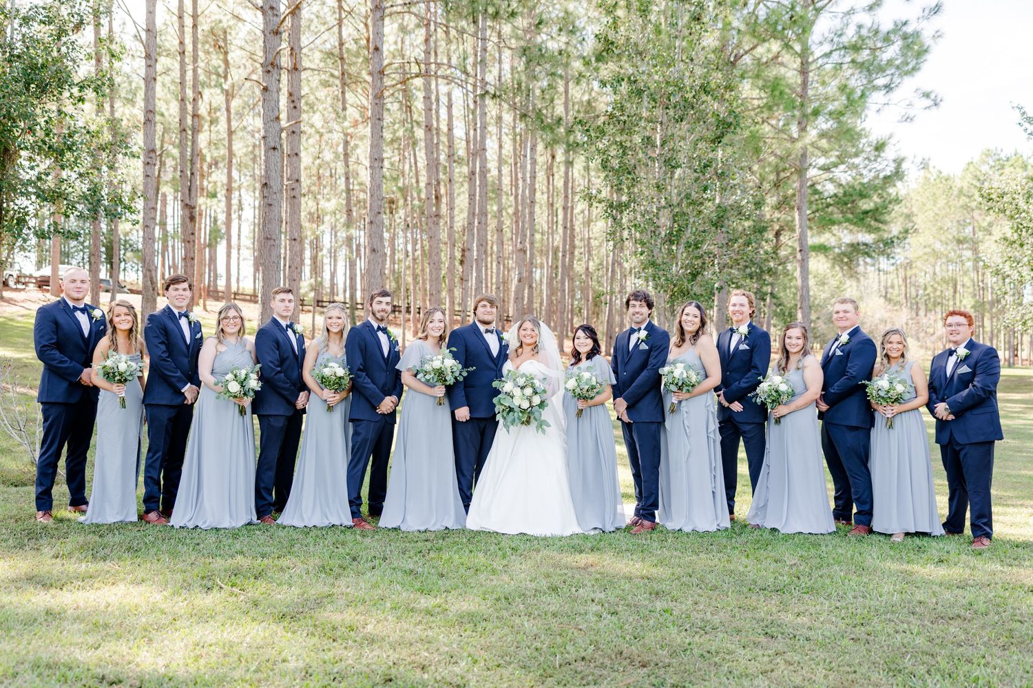 Wedding party members stand in formation outdoors with pine trees creating a natural backdrop.