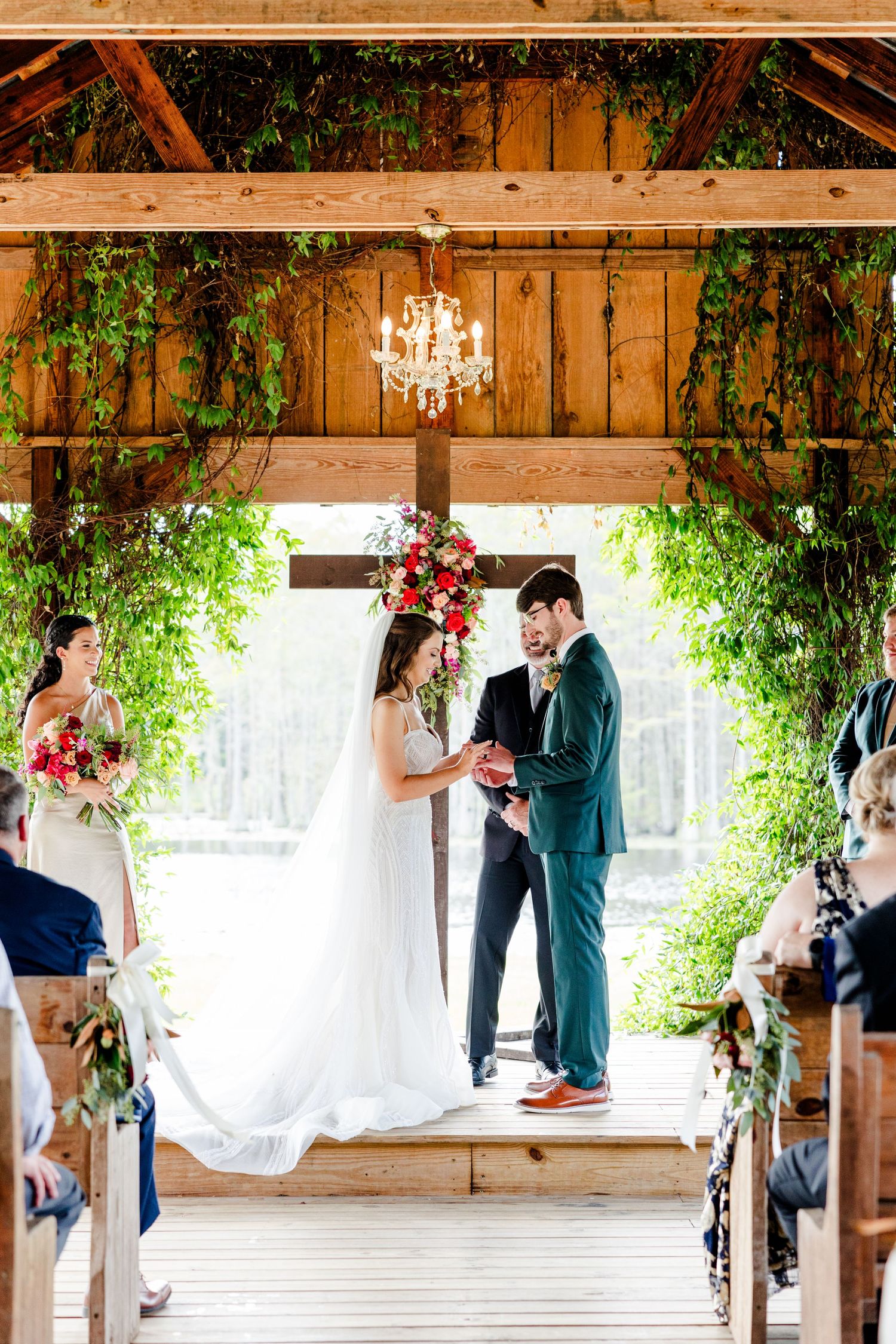 Couple exchanges vows at rustic barn wedding ceremony adorned with lush greenery, floral arch, and chandelier.