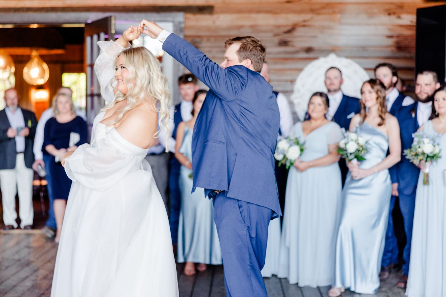 Wedding party in light blue bridesmaid dresses and navy suits dancing together at a rustic venue.
