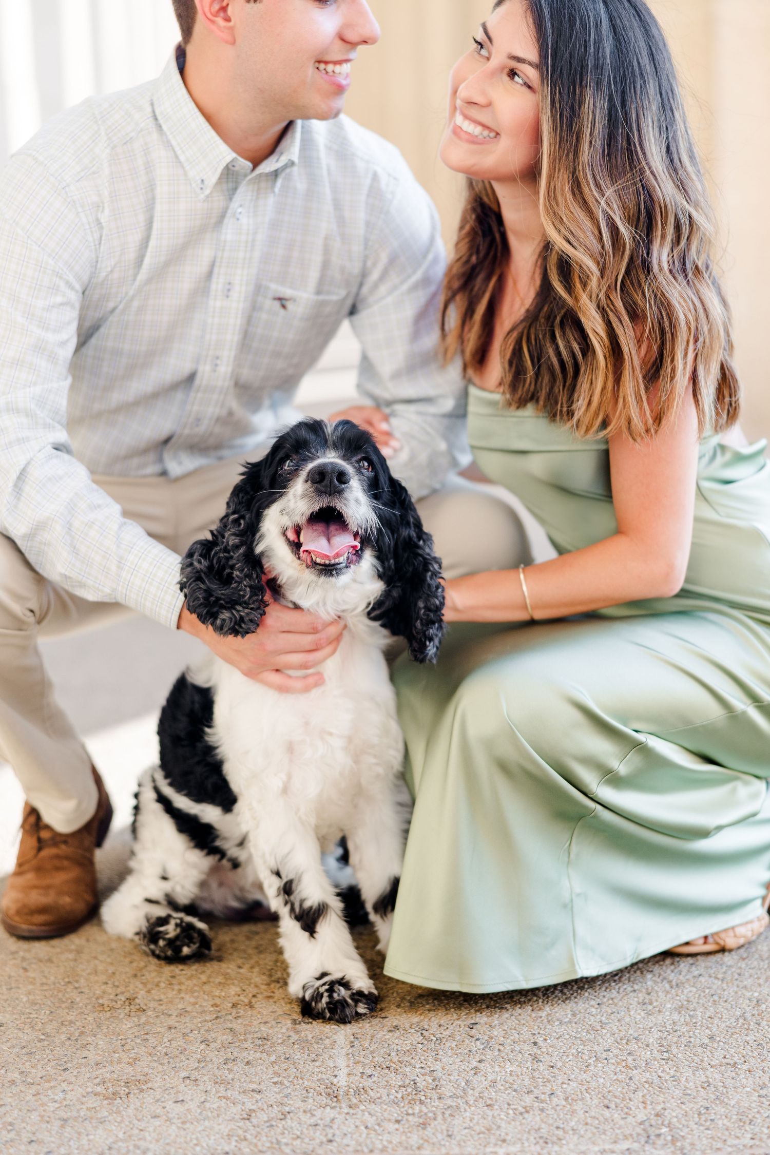 Smiling couple crouching with a black and white Cocker Spaniel dog, woman in sage green dress, man in light plaid shirt.